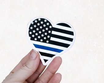 May include: A heart-shaped sticker with an American flag design and a blue stripe across the middle. The sticker is held in a hand.