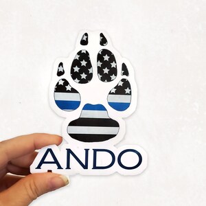 May include: A white sticker featuring a paw print design with a blue and black striped pattern and white stars. The word "ANDO" is printed in navy blue below the paw print.