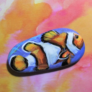 May include: Hand-painted rock art featuring a vibrant clownfish design. The fish, with its orange, white, and black markings, is set against a blue background. The artwork is on a smooth, oval-shaped stone, perfect for home decor.