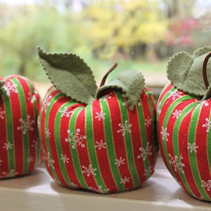 May include: Three red and green striped felt apples with white snowflake designs. The apples have green felt leaves on top.