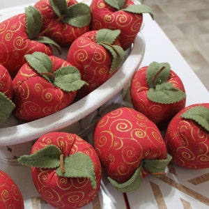 May include: A collection of red fabric apples with gold swirl patterns and green felt leaves. The apples are arranged in a white, shallow, oval-shaped bowl. The apples are a decorative item.