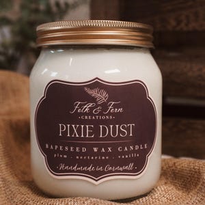 May include: A white jar candle with a gold lid and a dark burgundy label. The label reads "Pixie Dust" and lists the ingredients. The candle is on a burlap surface.