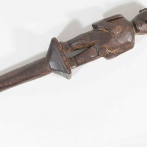 May include: A wooden carving of a standing figure with a long, thin handle. The figure is carved in a stylized, primitive style, with a large head and simple features. The handle is smooth and polished.
