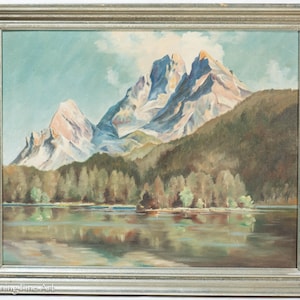 May include: Oil painting of a mountain landscape with a lake. The artwork features snow-capped mountains, a forest, and a reflective lake. The painting is framed in a silver-colored frame.