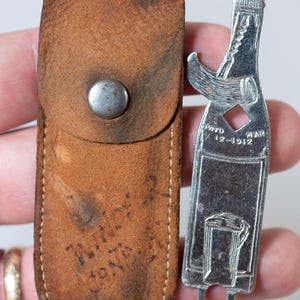 May include: A vintage bottle opener and leather case. The silver-colored opener is shaped like a bottle with a diamond-shaped cutout and the text "PATD MAR 12-1912". The brown leather case has a snap closure and shows signs of wear.
