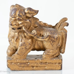 May include: A golden-colored, antique-style Foo Dog statue. The sculpture has a detailed, textured surface with visible wear and tear. It is mounted on a rectangular base, showcasing a traditional Asian art style.