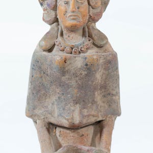 May include: A weathered terracotta sculpture of a figure with a detailed headdress and necklace. The figure is seated, holding a bowl-like object. The sculpture has a muted color palette with hints of blue and orange. The overall style suggests an ancient artifact.