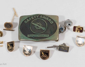 May include: A collection of vintage gold-tone pins and tie tacks featuring various logos, including "Martin Marietta", "Skylab", and "Antiair", along with a safety award buckle.