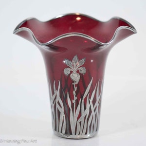 May include: A red glass vase with a ruffled top and silver-toned floral design. The vase features a stylized iris flower and leaves.