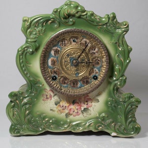 May include: A green porcelain mantle clock with ornate floral details and a painted floral design. The clock face has Roman numerals and a gold-toned finish.