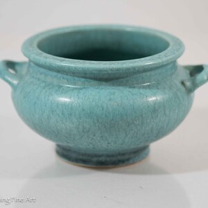 May include: A small, teal-colored ceramic pot with two handles. The pot has a speckled glaze and a rounded shape.