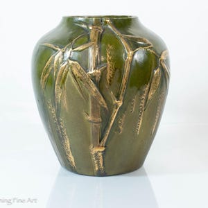 May include: A green ceramic vase with a gold bamboo design. The vase has a rounded shape and a glossy finish. The bamboo stalks and leaves are raised and detailed, creating a textured effect. The vase is a decorative item.