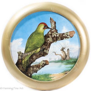 May include: A framed painting of a green woodpecker with a red head perched on a tree branch. The artwork is set against a backdrop of a blue sky with clouds and a landscape featuring a lake and trees. The frame is gold.
