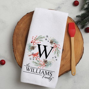 May include: White waffle-weave kitchen towel with a wreath design featuring a red cardinal, berries, and pinecones. The letter "W" is centered within the wreath, with "the WILLIAMS Family" printed below. A red spatula rests on a wooden cutting board.