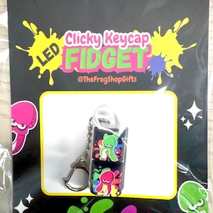 May include: A black and white keychain with a colorful LED light-up fidget toy. The toy features a cartoon octopus design and the text "Clicky Keycap LED Fidget @TheFrogShopGifts".