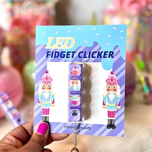 Könnte beinhalten: Ein lila und pinkfarbener LED-Fidget-Clicker mit einem Nussknacker-Design. Der Clicker ist an einem silbernen Schlüsselanhänger befestigt. Die Verpackung zeigt den Text "LED Fidget Clicker" und "@TheFrashopGifts".