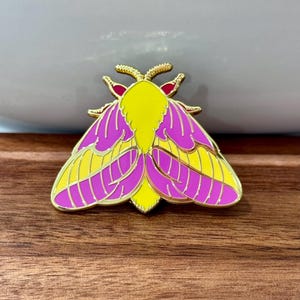 Puede incluir: Un pin de esmalte dorado con una polilla amarilla y rosa con antenas rojas.
