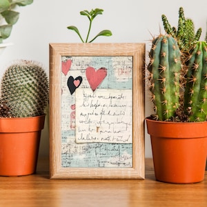 May include: A framed print with a vintage aesthetic featuring a collage of handwritten text, paper scraps, and pink and black hearts. The print is displayed on a wooden table with potted cacti on either side.
