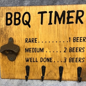 May include: Wooden BBQ timer with a bottle opener and hooks. The timer has a black painted inscription that reads "BBQ TIMER" and "RARE... 1 BEER", "MEDIUM... 2 BEERS", "WELL DONE... 3 BEERS".