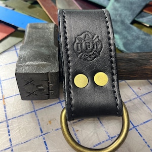 May include: Black leather axe loop with a brass ring and two brass rivets. The loop has an embossed firefighter emblem with the letters "I.F.F."