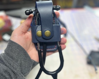 May include: A dark blue leather stethoscope holder with two gold-toned buttons. The holder is designed to hold a stethoscope with a black tube and a black and silver head.