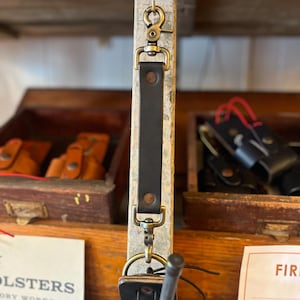 May include: A black leather key chain with a brass swivel hook and a brass snap hook. The key chain is hanging from a wooden shelf.