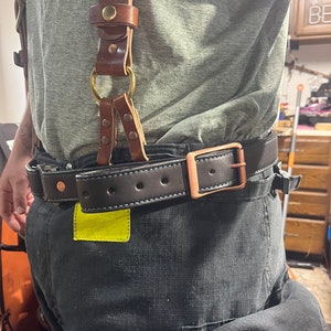 May include: A brown leather suspender with brass hardware and a black leather belt with a copper buckle. The belt is worn over a pair of black work pants.
