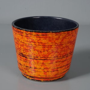 Könnte beinhalten: Ein Keramik-Pflanztopf mit einer lebendigen orangefarbenen und schwarzen strukturierten Außenseite. Der Innenraum ist ein tiefes Marineblau. Der Topf hat eine zylindrische Form, geeignet für verschiedene Pflanzen.