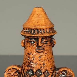 Peut inclure: Une figurine en céramique de couleur terre cuite avec un sommet conique, présentant un visage stylisé et des oreilles circulaires saillantes. Le corps est orné de motifs géométriques incisés, incluant des carrés et des triangles, en finition mate.
