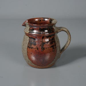 Puede incluir: Una jarra de cerámica hecha a mano con pico y asa. La jarra presenta un exterior moteado en tonos beige y marrón con un centro rojizo-marrón. El borde y el interior son de color marrón oscuro.