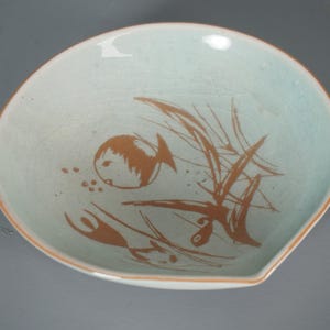 Pode incluir: Um prato de cerâmica azul claro com um desenho de peixe e plantas aquáticas marrons. O prato tem uma forma única com uma borda ligeiramente elevada e uma borda marrom. A obra de arte é uma ilustração simples, mas elegante.