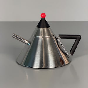 Könnte beinhalten: Eine konische Teekanne aus Edelstahl mit polierter Oberfläche. Sie verfügt über einen schwarzen, dreieckigen Deckel mit einer kleinen roten Kugel an der Spitze, einen geraden Metallauslauf und einen schwarzen, eckigen Griff an der Seite.