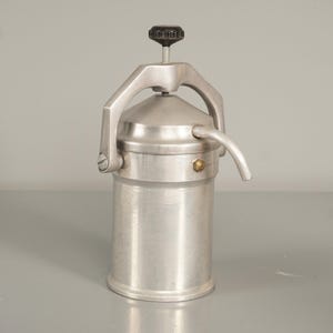Könnte beinhalten: Ein Vintage-Espressokocher aus gebürstetem Aluminium. Der zylindrische Körper hat einen gebogenen Ausguss und einen Griff, der sich über die Oberseite wölbt. Oben auf dem Gerät befindet sich ein schwarzer Knopf.