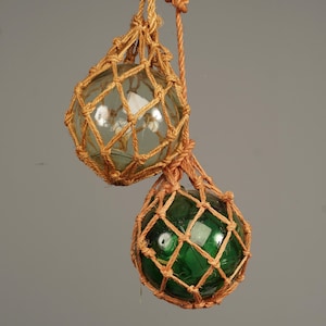 Könnte beinhalten: Zwei Glas-Fischerschwimmer, einer klar und einer grün, aufgehängt an einem gewebten, hellbraunen Seilnetz. Die Schwimmer sind rund und haben eine glänzende Oberfläche. Das Seil ist verknotet, um die Schwimmer zu halten.