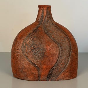 Könnte beinhalten: Eine terrakottafarbene Vase mit einem abgerundeten, abgeflachten Körper und einem schmalen Hals. Die Oberfläche weist eine raue, strukturierte Textur mit dunkelbraunen, geschwungenen, vertieften Rillen auf, die Tiefe erzeugen.