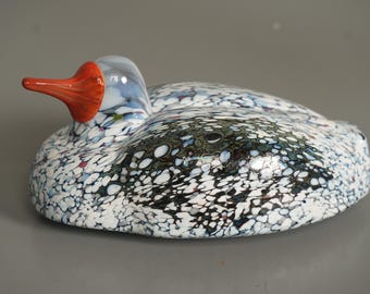 May include: A decorative glass duck figurine. The duck has a speckled pattern in white, black, and blue. It features a red-orange beak and a clear head with blue accents. The figurine is a unique home decor item.