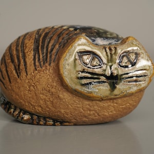 May include: A ceramic cat figurine in a seated position. The cat has a tan body with dark brown stripes and a light green and tan face. The eyes are detailed with black lines, and the overall design is rounded and stylized.