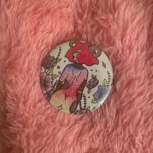 May include: A white button featuring a colourful illustration of a person with red hair and horns, wearing a gradient shirt and black fishnet stockings. The person is surrounded by flowers and stars.
