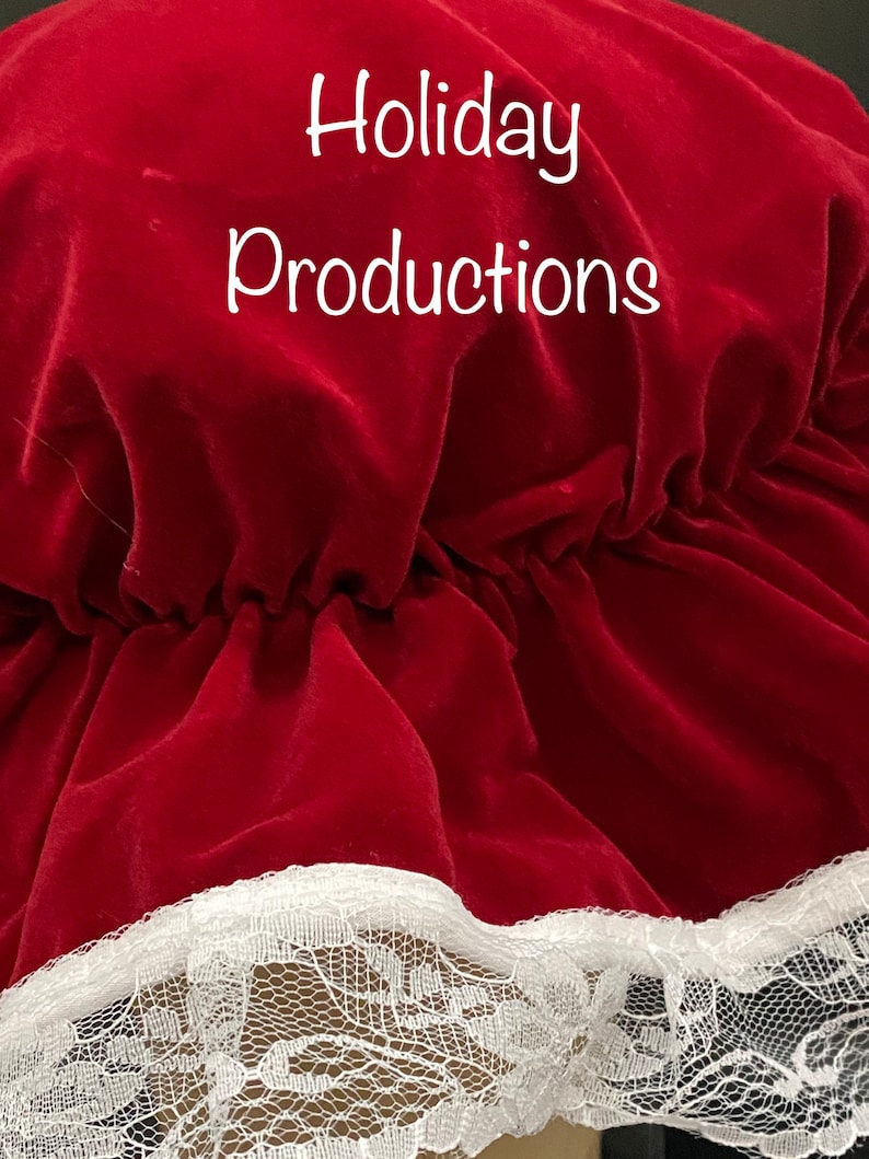 Pu&ograve; includere: Primo piano di un cappello di velluto rosso con finiture in pizzo bianco. Il testo "Holiday Productions" &egrave; scritto in bianco sulla parte superiore del cappello. Il cappello ha un design a balze.