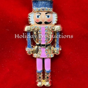 May include: A nutcracker brooch featuring a blue glitter hat, gold-tone metal accents, and pink enamel details. The figure is adorned with clear iridescent rhinestones along the jacket and hat brim. The text Holiday Productions is overlaid.