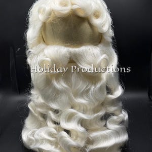 May include: A white wig and beard set featuring long, wavy hair with curled ends and a matching mustache. The hair is styled with a center part and voluminous curls. The text "Holiday Productions" is printed in white across the center of the beard.
