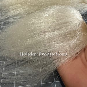 May include: Close-up of light blonde faux fur. The fur is thick and has a shiny appearance. The text "Holiday Productions" is visible. The fur is on a cutting mat with inch measurements.