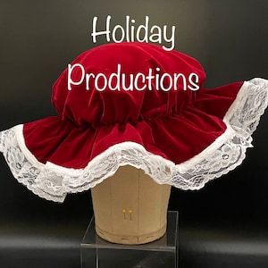 Pu&ograve; includere: Un cappello di velluto rosso con un'ampia tesa, rifinito con pizzo bianco. Il testo "Holiday Productions" &egrave; visualizzato nella parte superiore dell'immagine. Il cappello &egrave; esposto su una testa di manichino.