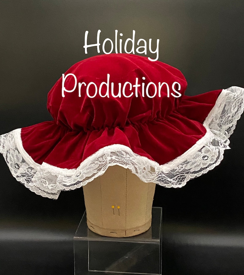 Pu&ograve; includere: Un cappello di velluto rosso con un'ampia tesa, rifinito con pizzo bianco. Il testo "Holiday Productions" &egrave; visualizzato nella parte superiore dell'immagine. Il cappello &egrave; esposto su una testa di manichino.