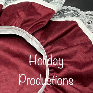 Pu&ograve; includere: Primo piano di tessuto bordeaux con bordatura in pizzo bianco e fascia elastica. Il testo "Holiday Productions" &egrave; stampato in bianco. Il tessuto ha una leggera lucentezza.