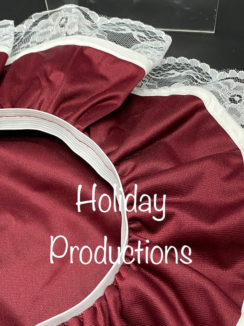 Pu&ograve; includere: Primo piano di tessuto bordeaux con bordatura in pizzo bianco e fascia elastica. Il testo "Holiday Productions" &egrave; stampato in bianco. Il tessuto ha una leggera lucentezza.
