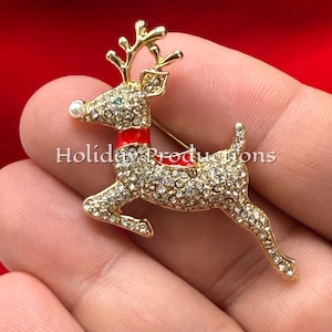 May include: A gold-toned reindeer brooch featuring clear rhinestones covering the body, a bright red enamel collar, and a small white pearl-like bead for the nose. The text Holiday Productions is overlaid across the center of the reindeer figure.