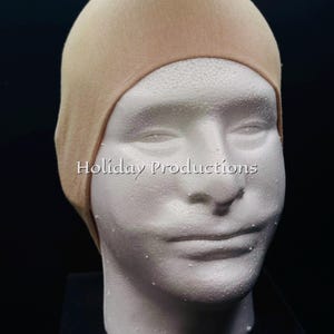 May include: A beige wig cap is displayed on a white foam mannequin head. The cap is made of a stretchy fabric and covers the head, ears, and hairline. The text "Holiday Productions" is visible on the image.