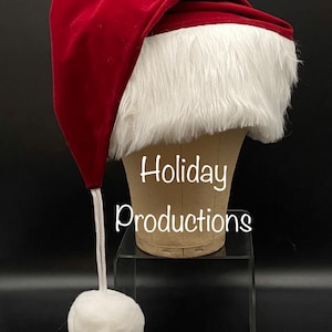 May include: A red velvet Santa hat with a white faux fur trim and a white pom-pom on the end of the long cord. The hat is on a mannequin head with the text "Holiday Productions" written below.