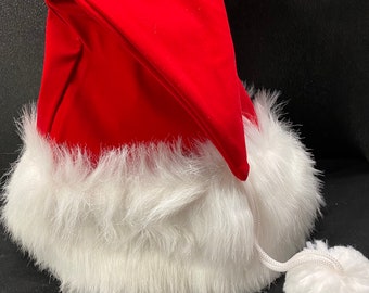 Puede incluir: Un gorro de Papá Noel rojo con un borde de piel sintética blanca y un pompón blanco en el extremo de la cuerda del gorro.